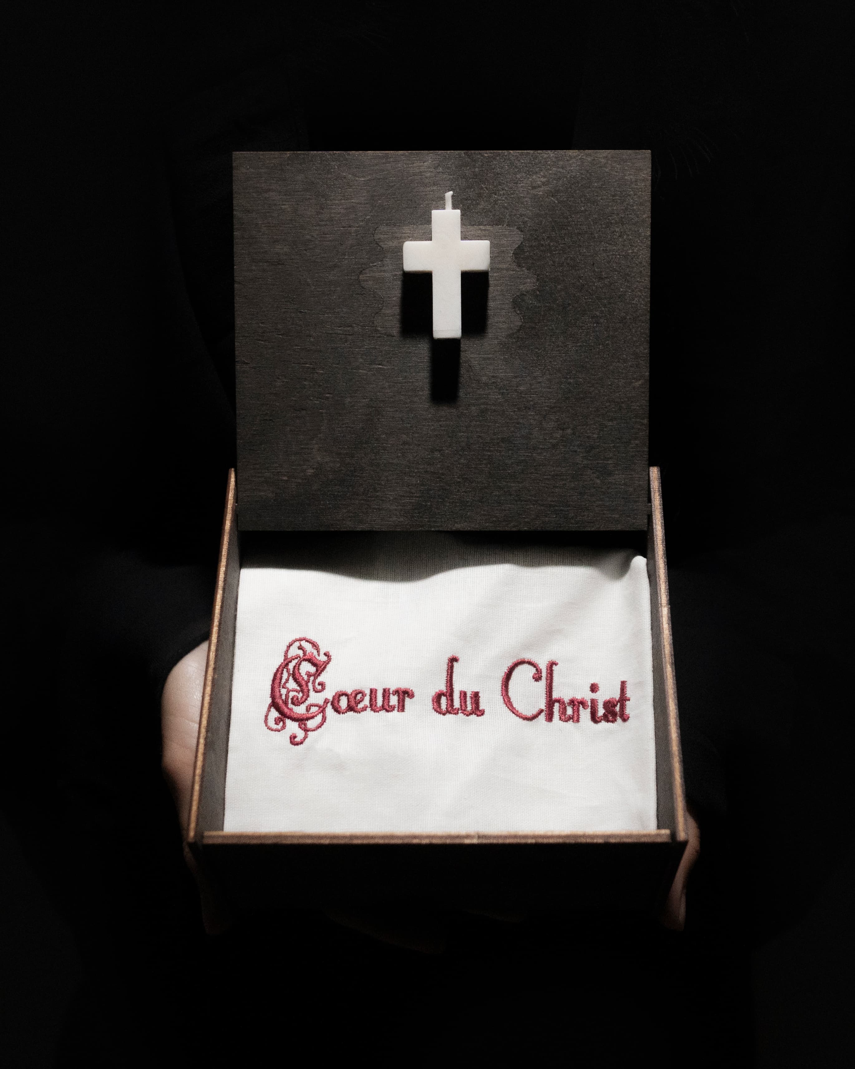 Open dark wooden box held in hands, showing a white cross candle on the lid and embroidered fabric inside