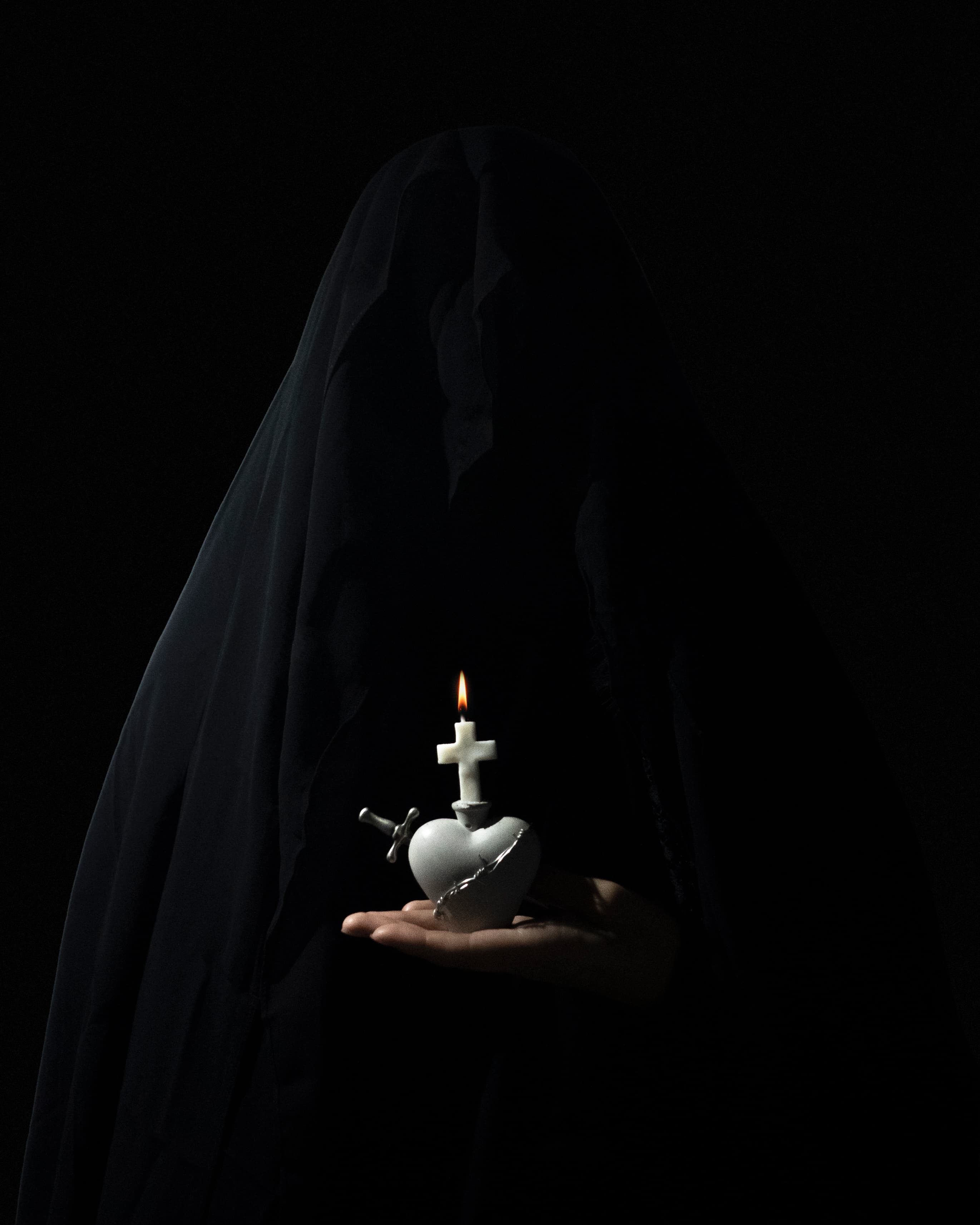 A figure in a black veil holds a white stone heart candle with a cross wick and a silver sword on a dark background