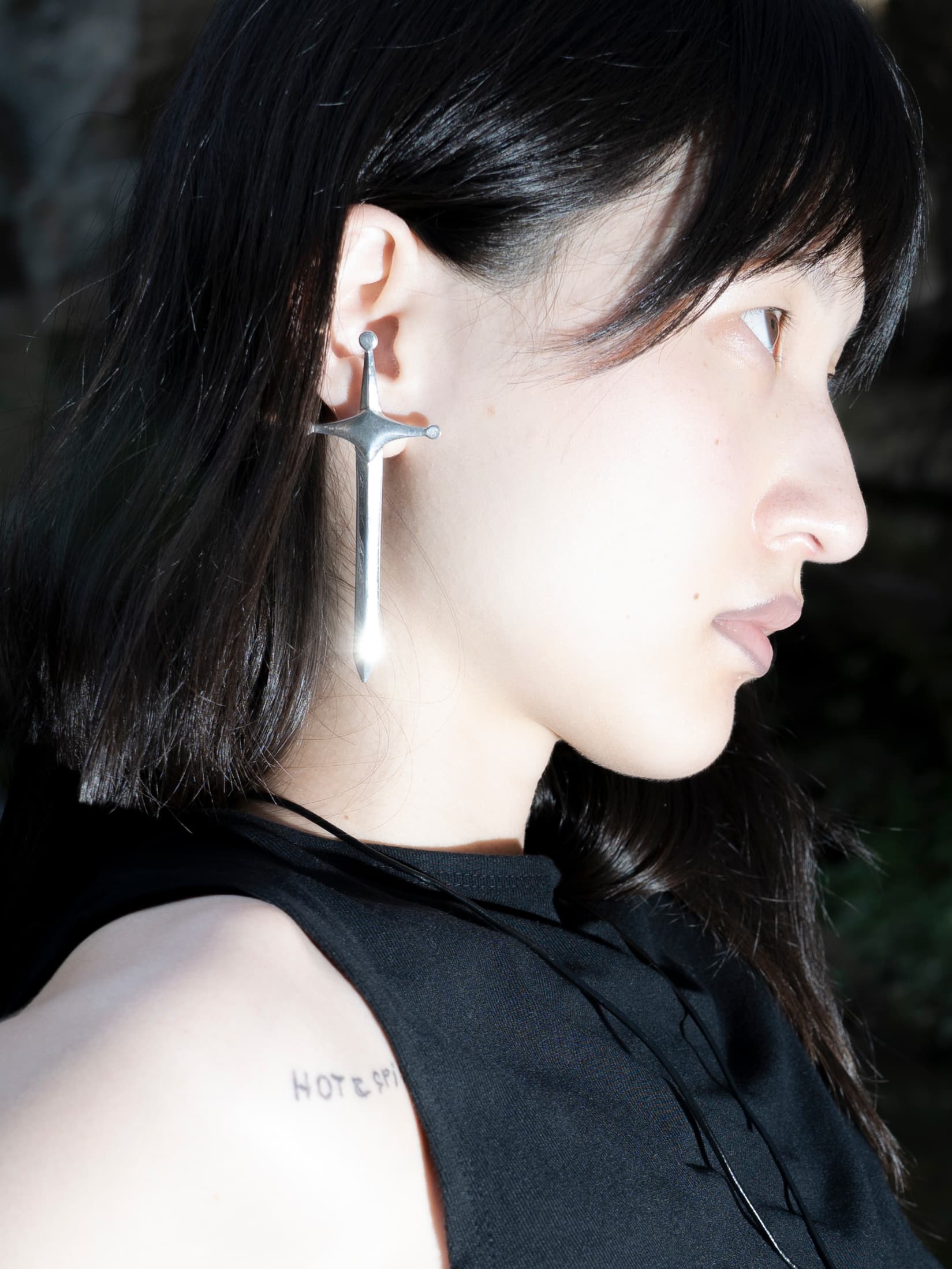 Profile portrait against moss-shaded stone: model with a long silver dagger earring, black tank, dark hair falling loose.