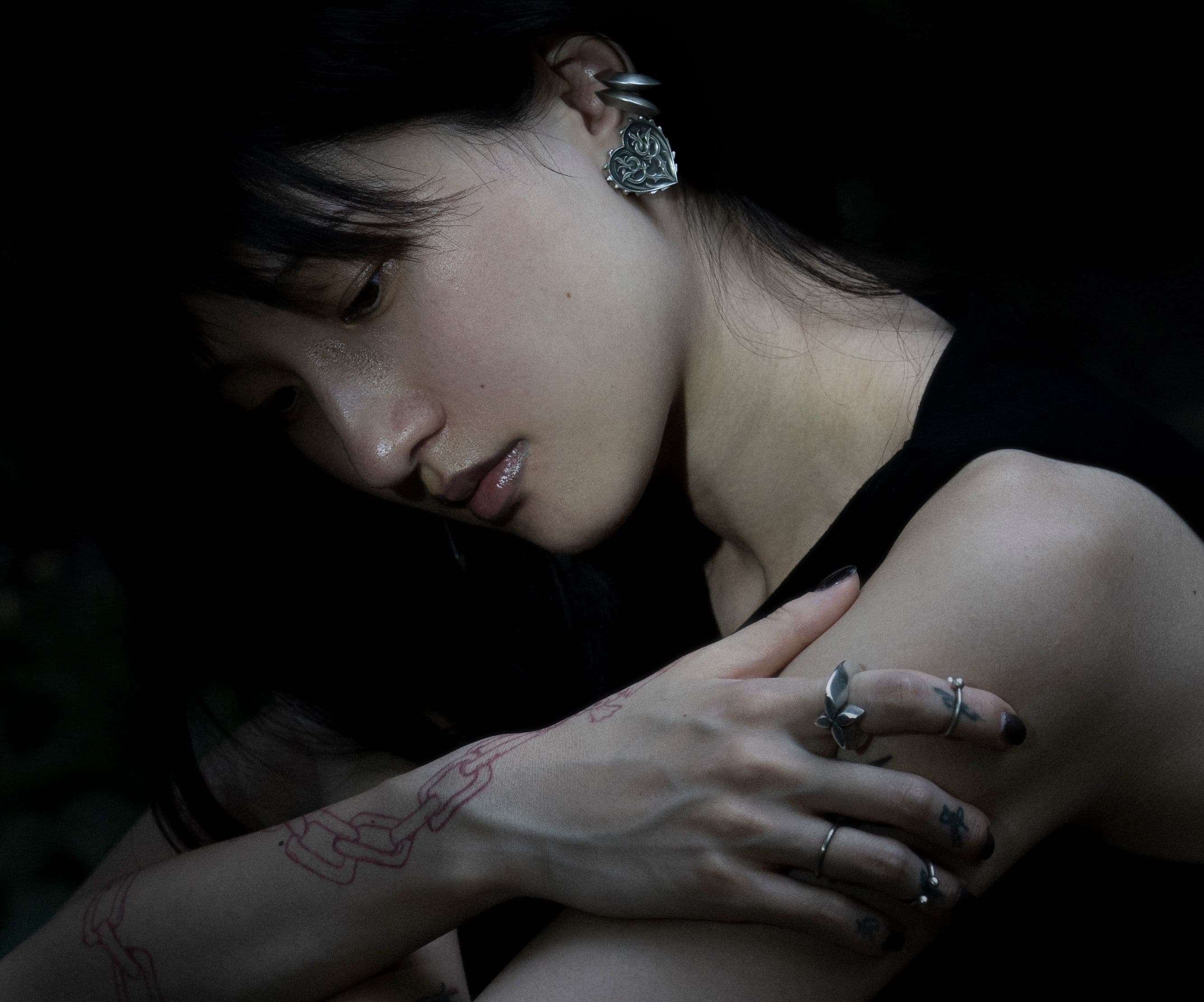 Portrait in low light: model wearing a heart-shaped silver ear cuff and floral silver rings, a chain traced in red across the forearm.