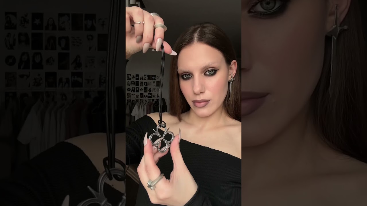 A model with dark makeup holds a silver quatrefoil pendant on a black cord, wearing matching silver rings